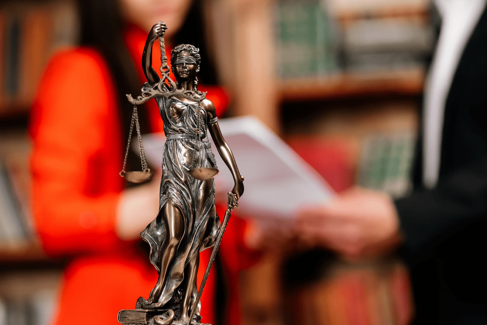 A professional photo shows a bronze statue of Lady Justice in the foreground. She is blindfolded, holding a set of scales in one hand and a sword in the other. In the blurred background, a person in a red blazer is talking to a person in a black suit, and they are both holding papers, suggesting they are discussing a legal matter. A bookshelf is also visible in the background.
